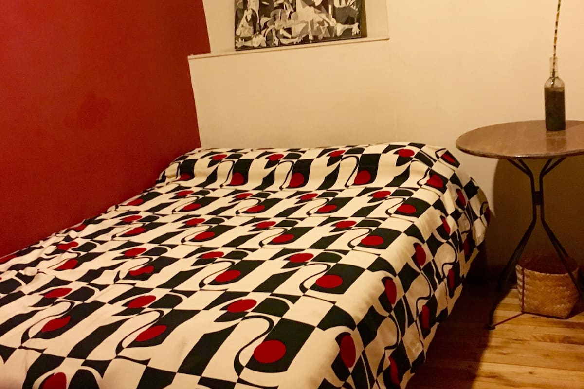 A bedroom with a patterned bedspread, a small round table, a potted plant, and a wall-mounted artwork.