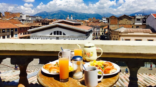 Un balcone con un vassoio per la colazione, che include croissant, succo d'arancia e un teiera, con vista su un panorama urbano che spazia tra edifici storici e montagne sullo sfondo.