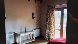 Une pièce avec un plafond en bois, un bureau blanc avec une lampe, une fenêtre ornée de rideaux à motifs floraux et deux lits.