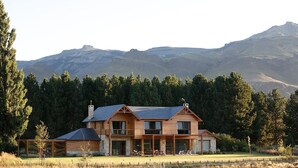 Une maison en bois à deux étages, dotée d’un grand porche à l’avant et de nombreuses fenêtres, entourée d’une forêt et avec des montagnes en arrière-plan.