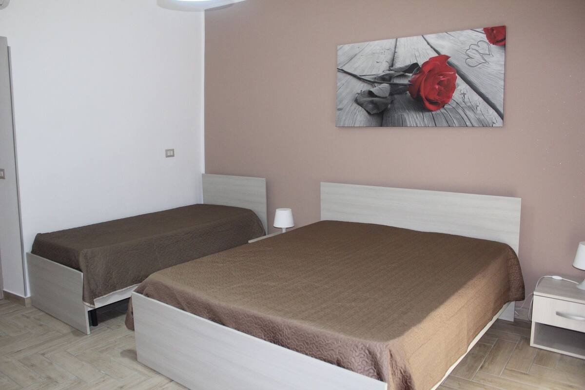 Una habitación con dos camas individuales, un cabecero blanco, una mesita de noche con lámpara y un cuadro colgado en la pared de una rosa roja.