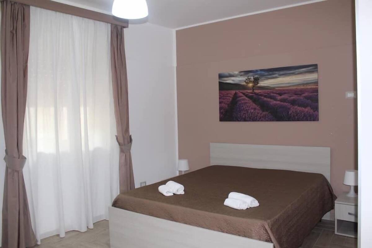 Un dormitorio con cama, cortinas blancas, un cuadro de un paisaje y una mesita de noche con lámpara.