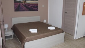 Una cama bien hecha con cubrecama marrón, dos toallas blancas, una mesita de noche con lámpara y un cuadro de campos de lavanda en la pared.