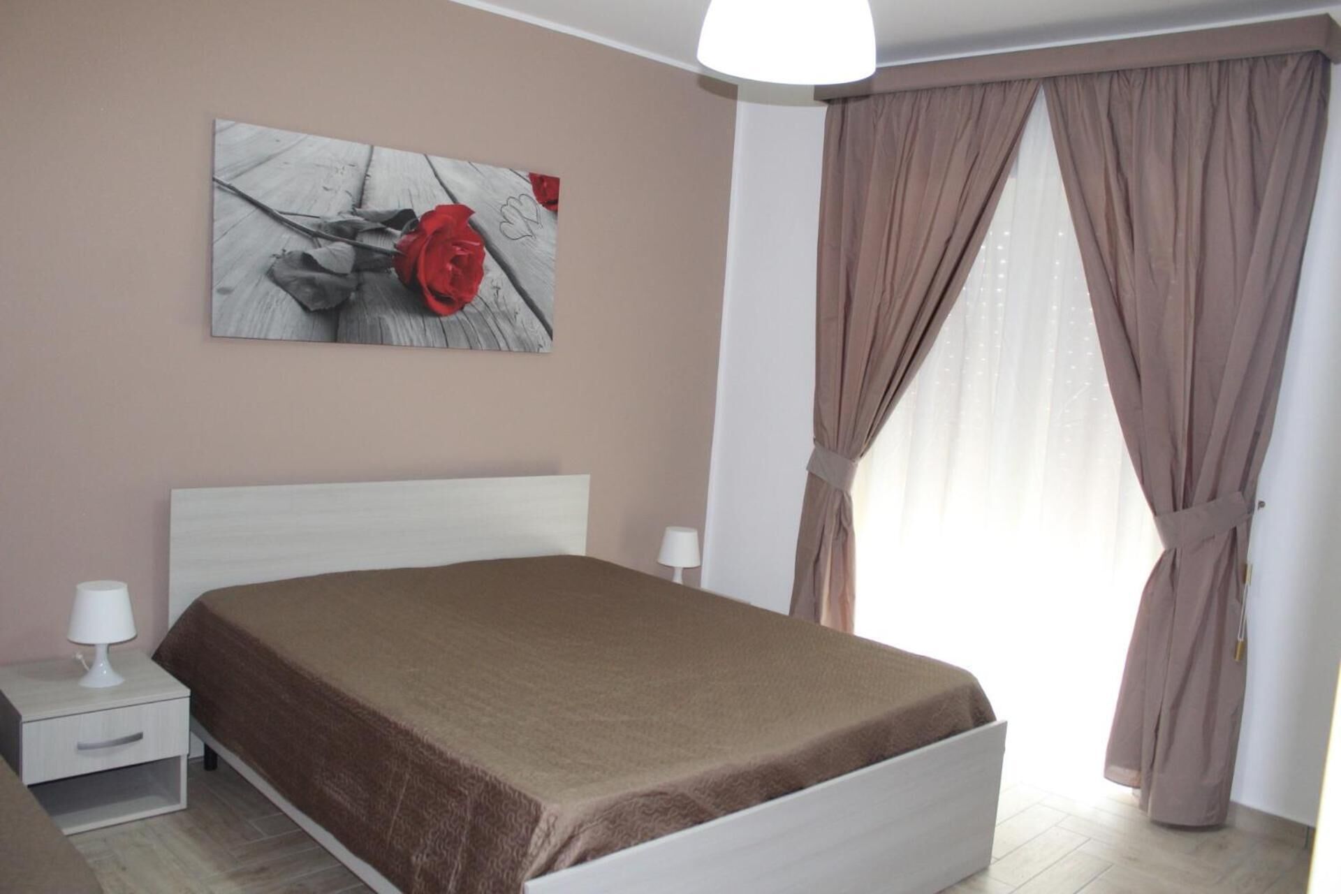 A bedroom with a bed, a nightstand, a lamp, a painting of a rose, and a window with curtains.