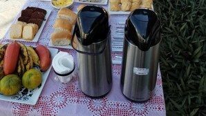 Un área de comedor al aire libre con una mesa dispuesta con diversos alimentos, entre panes, pasteles y frutas, y dos termos.