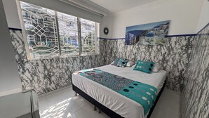 Un dormitorio con cama, ventana con rejas, pintura de una escena costera y pared con estampado de mármol.