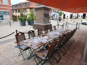 Ein Essbereich im Freien mit einem langen Tisch, Stühlen und einem roten Sonnenschirm.