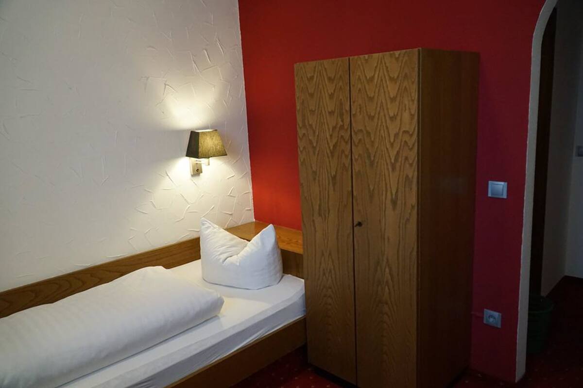 Una cama individual con ropa de cama blanca, un armario de madera y una lámpara fijada a la pared.