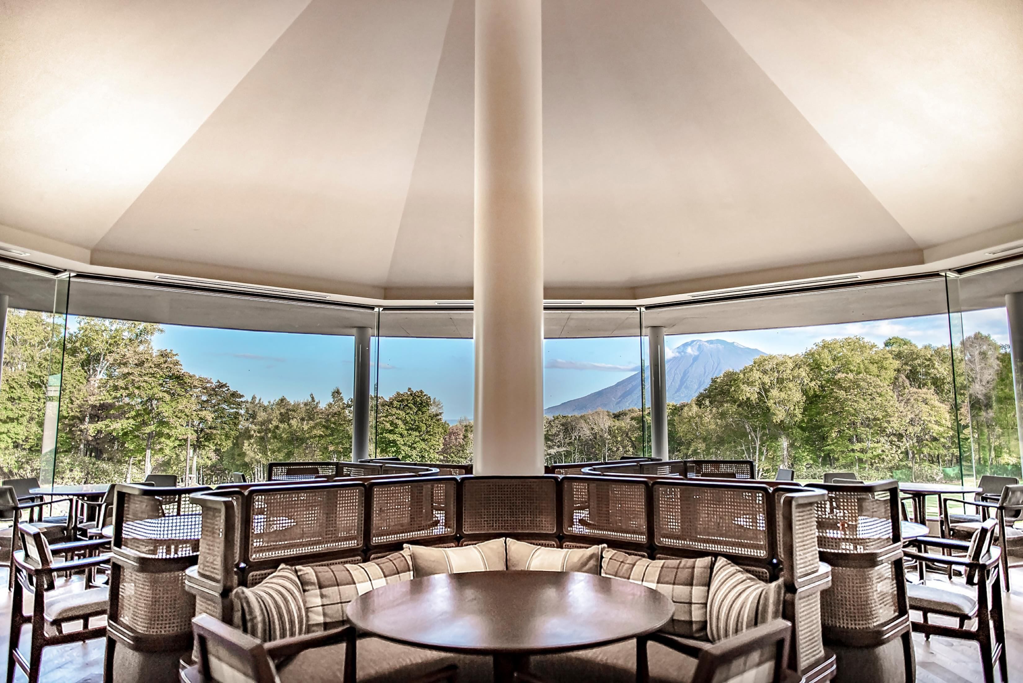 A round table with cushioned seating and wicker backrests in a covered outdoor area with large windows offering a mountain view.