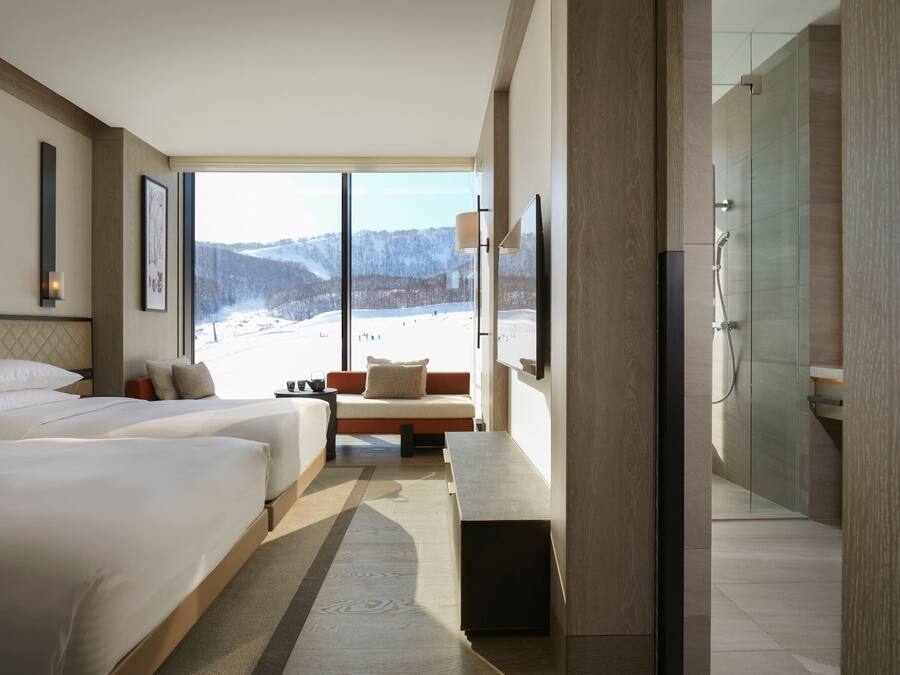 A modern hotel room with two beds, a seating area, a large window with a snowy mountain view, and a glass-enclosed shower area.