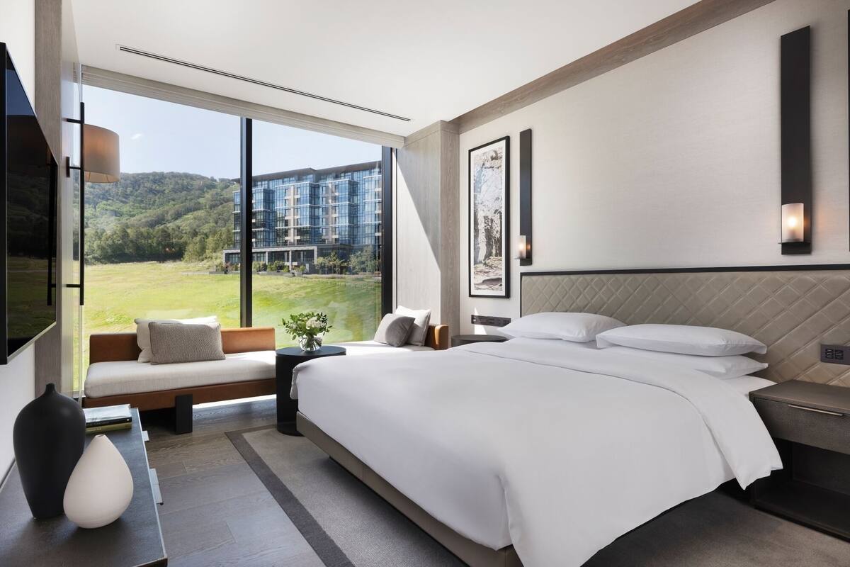 Ein modernes Hotelzimmer mit einem großen Bett, einer Sitzecke am Fenster und Blick auf ein Gebäude und Grünflächen.