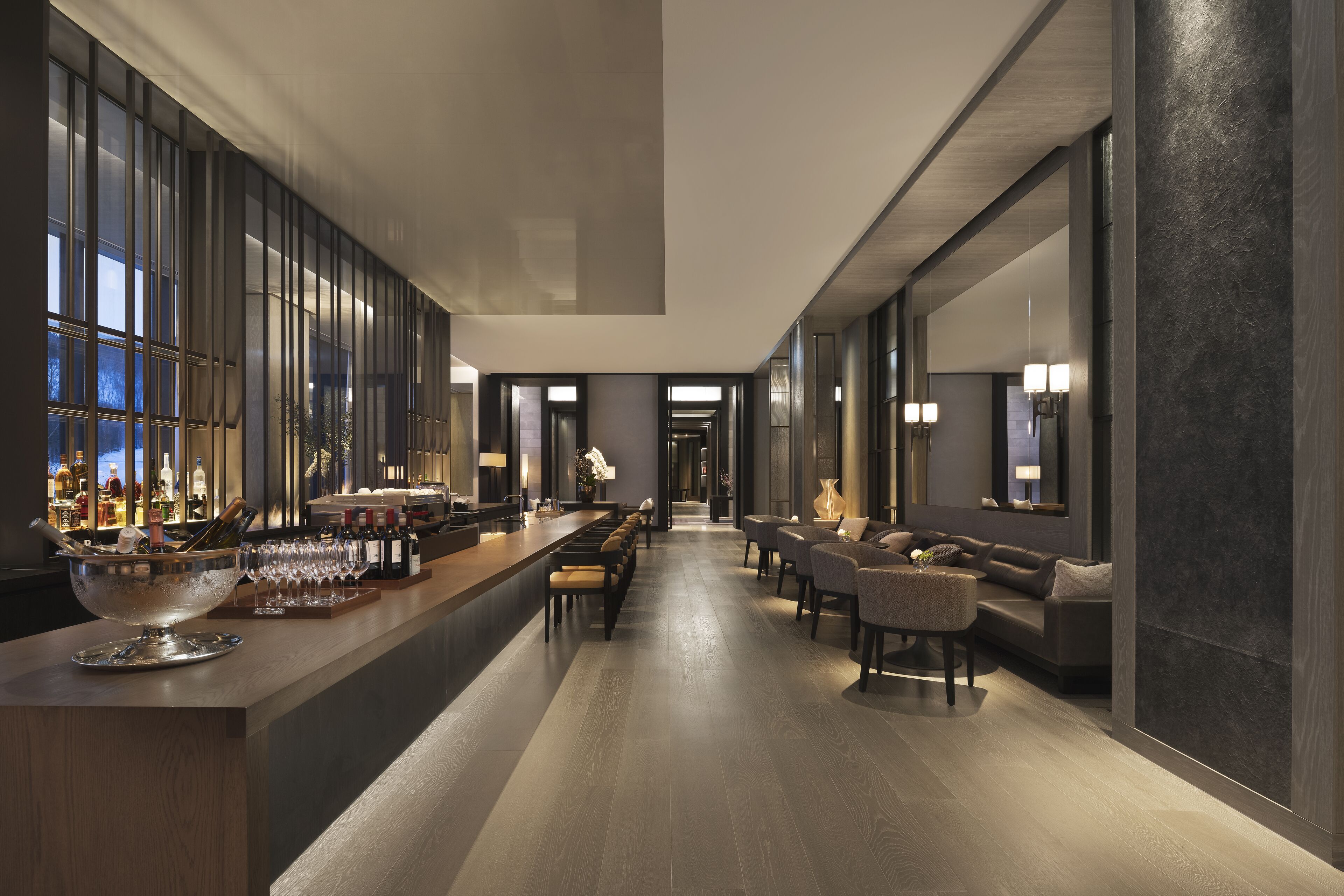 A modern bar area with a long counter, bar stools, and a large bowl of ice. There are bottles and glasses on the counter, and the room has a wooden floor and dark walls.