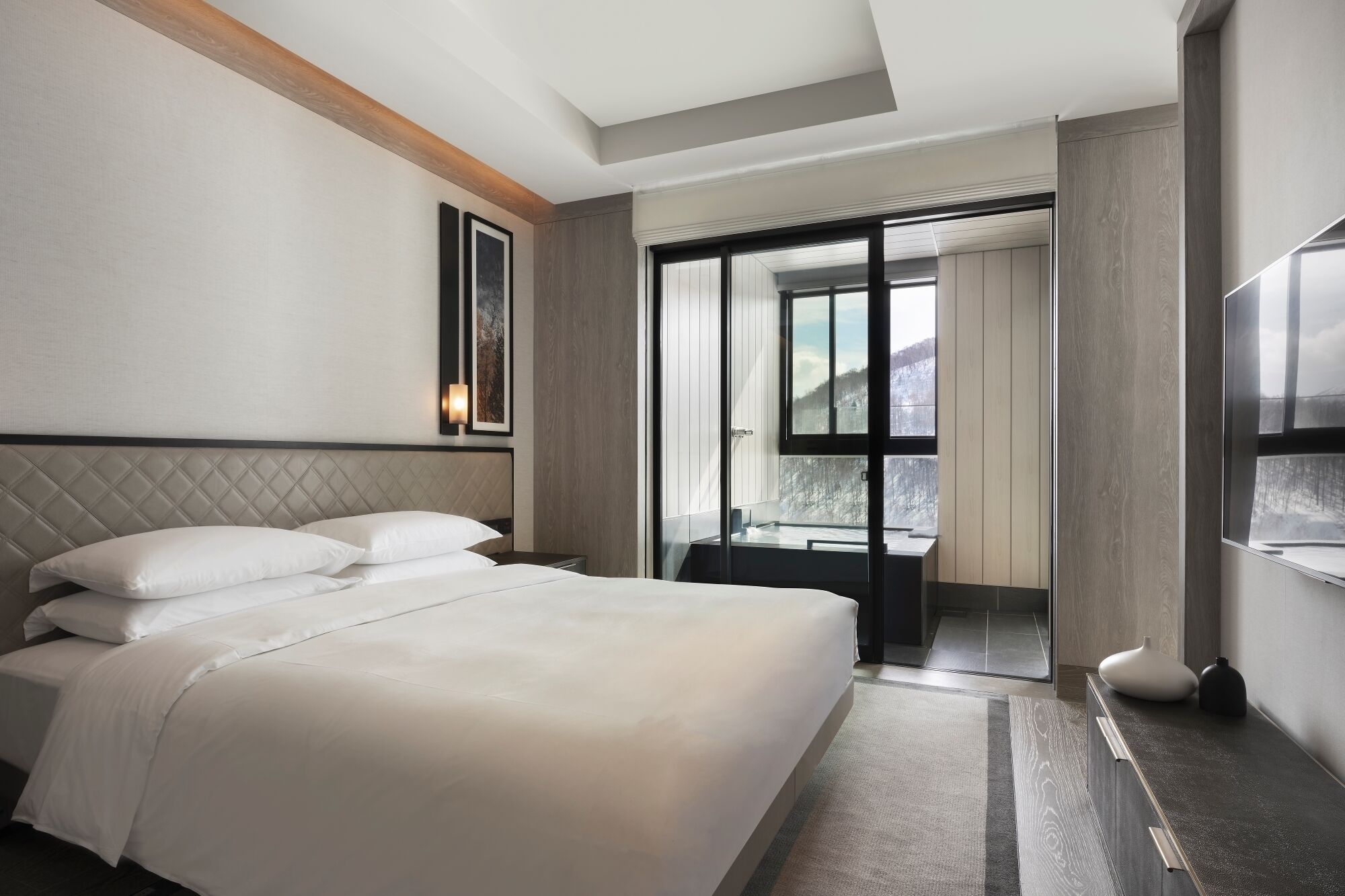 Una habitación de hotel moderna con una cama grande, una puerta de vidrio que da a un balcón y vistas a un paisaje nevado.