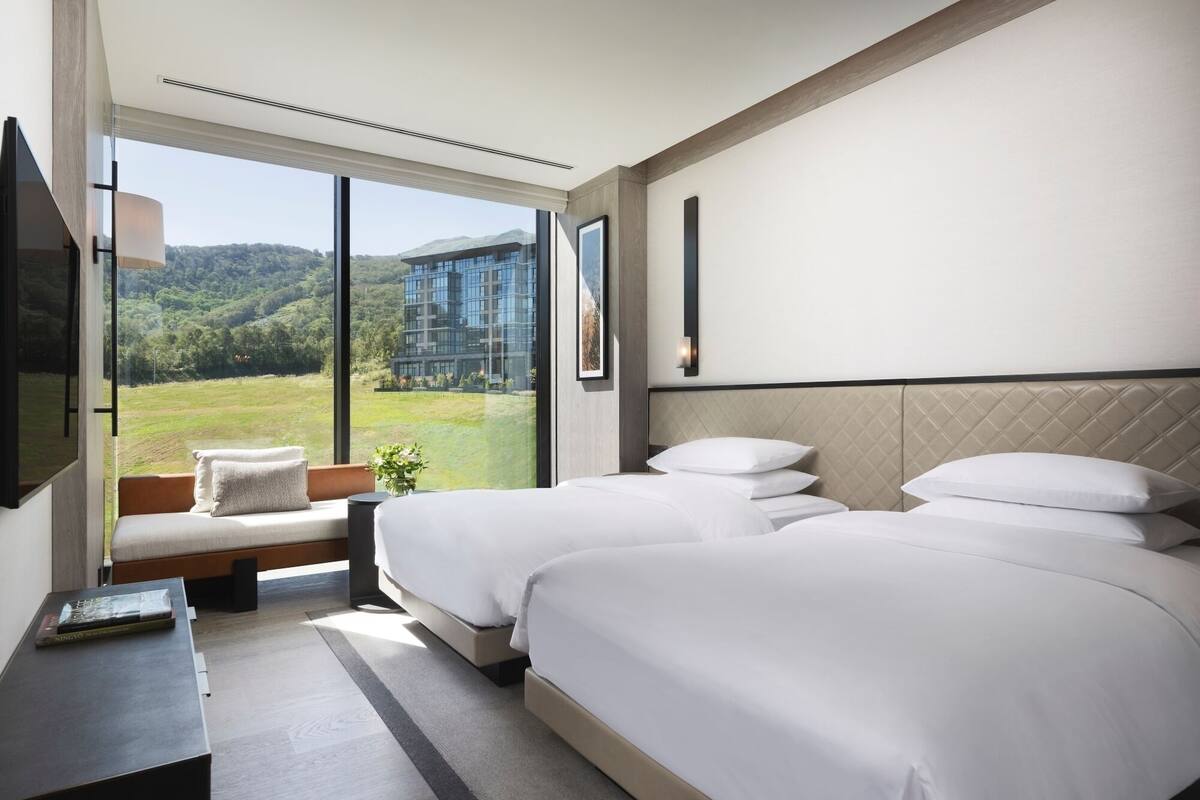 Ein Hotelzimmer mit zwei Betten, einem großen Fenster mit Blick auf ein Gebäude und Hügel, einem Nachttisch mit einem Buch und einem am Wand montierten Fernseher.