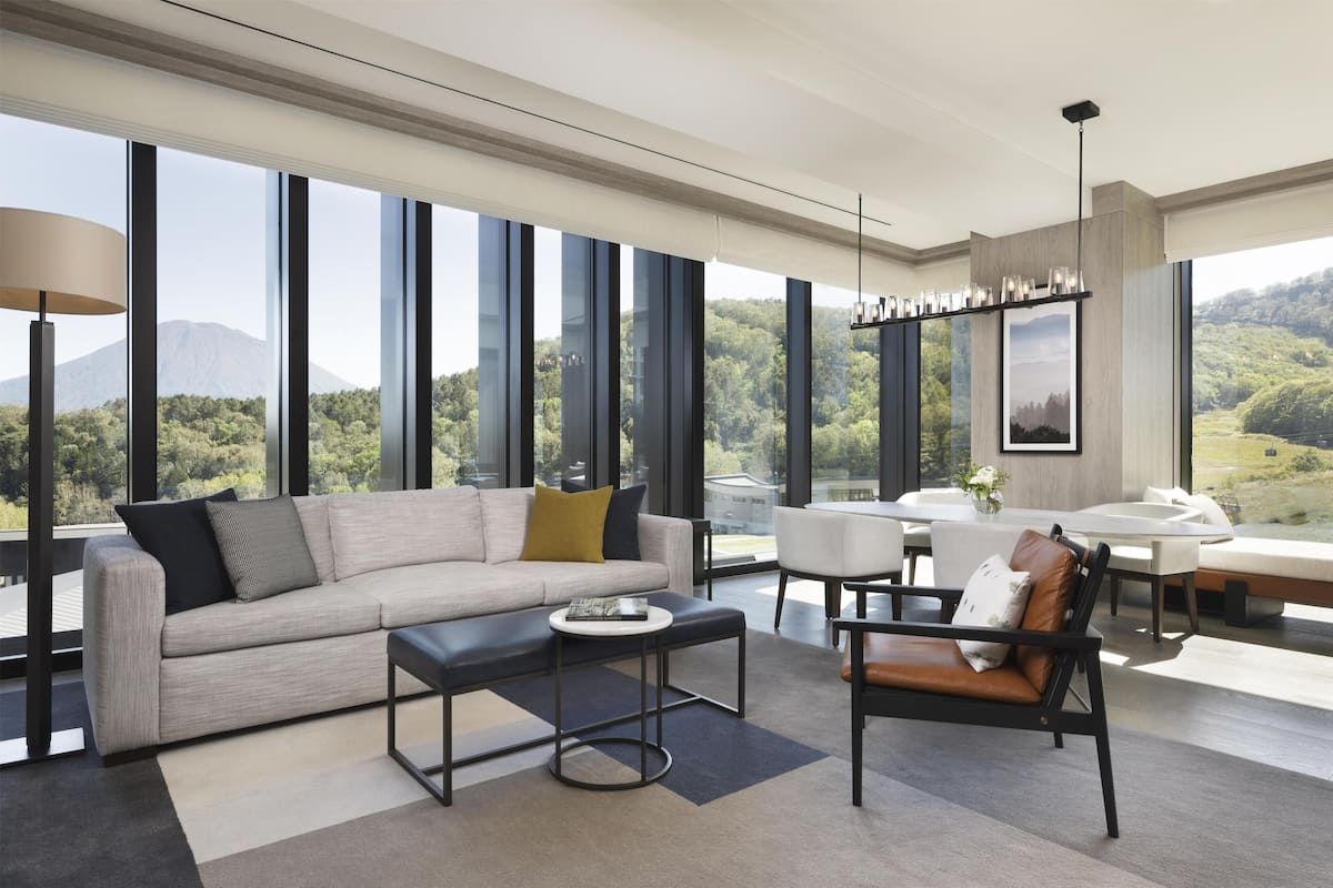 Ein modernes Wohnzimmer mit einer Couch, einem Couchtisch und Sesseln, großen Fenstern mit Bergblick und einem Essbereich mit Tisch und Stühlen.