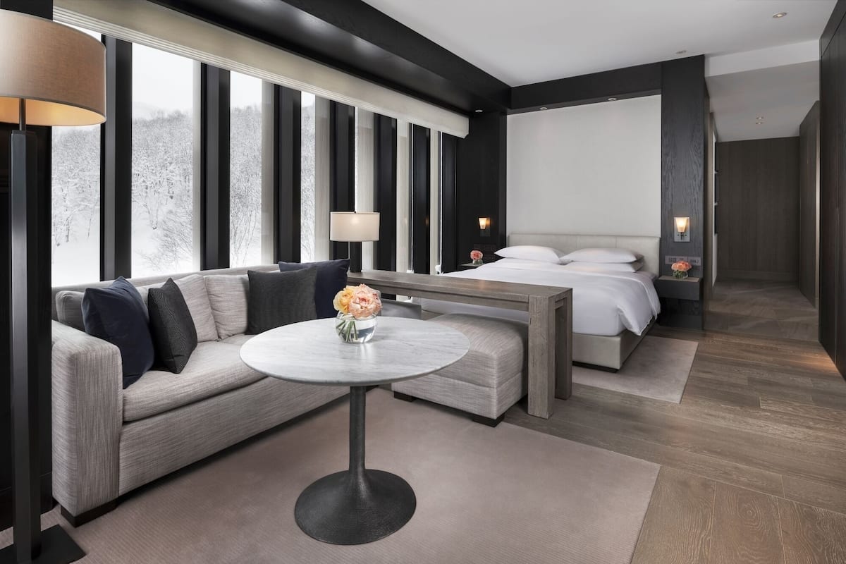 Ein modernes Hotelzimmer mit einer Couch, einem runden Tisch mit Blumenvase, einem Bett mit Nachttisch und einem großen Fenster mit Blick auf Bäume.