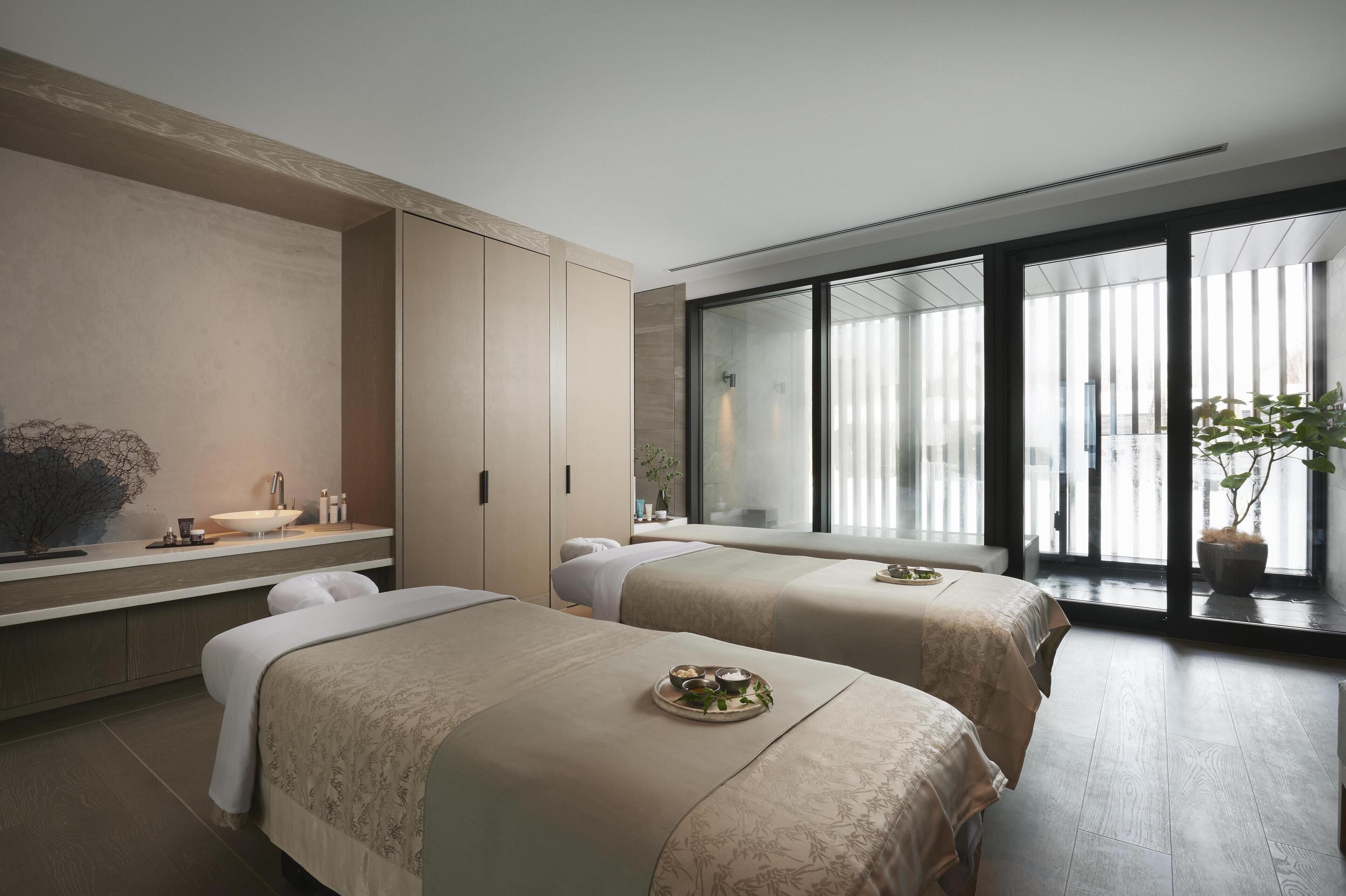 Een moderne hotelkamer met twee bedden, een groot raam, een wastafel en een plant.