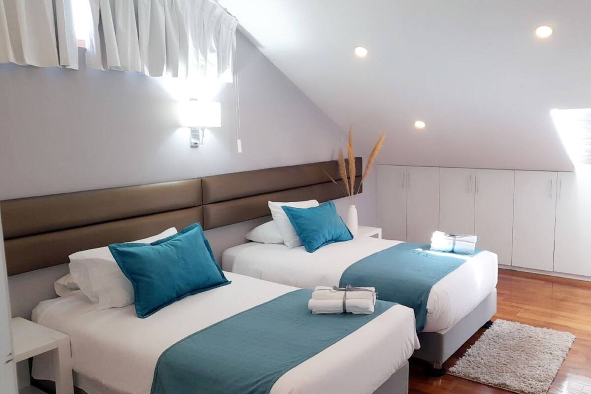 A modern bedroom with two beds, each with teal and white bedding, a wooden headboard, and a white nightstand.