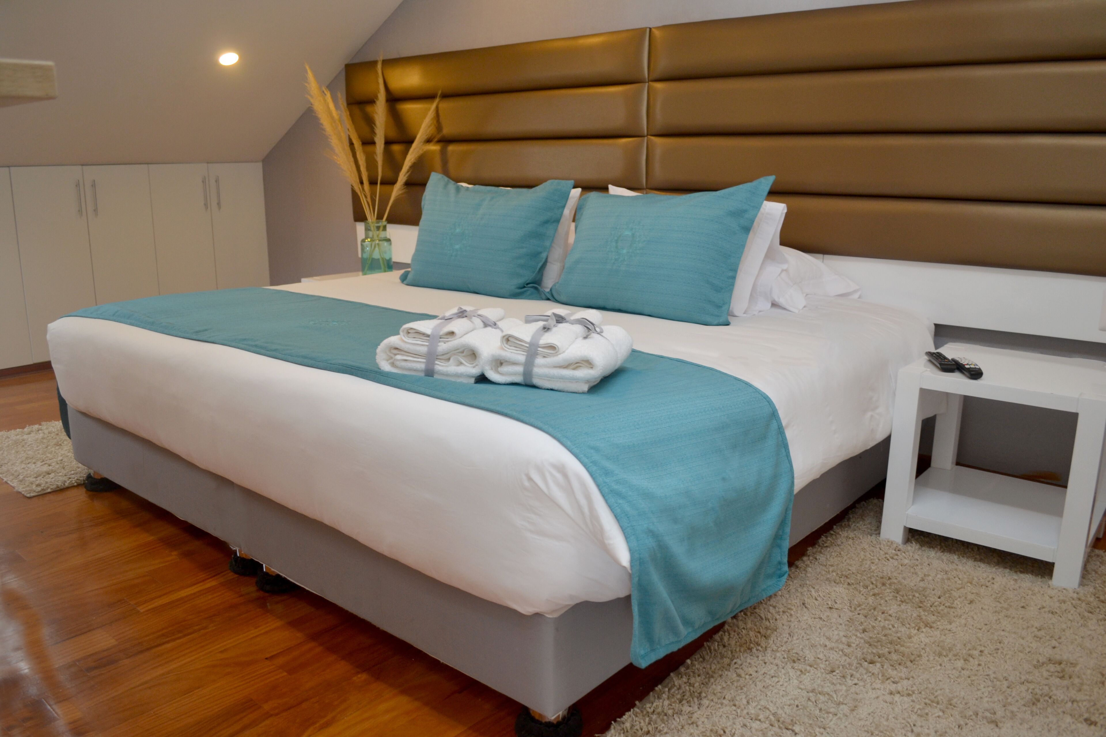 A neatly made bed with a teal blanket and pillows, a white bedside table with a remote control, and a wooden floor.
