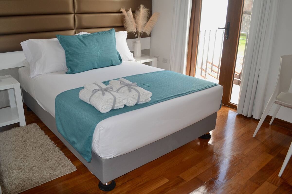 A neatly made bed with a teal throw blanket and white pillows, a wooden headboard, and a white nightstand with a vase of pampas grass.