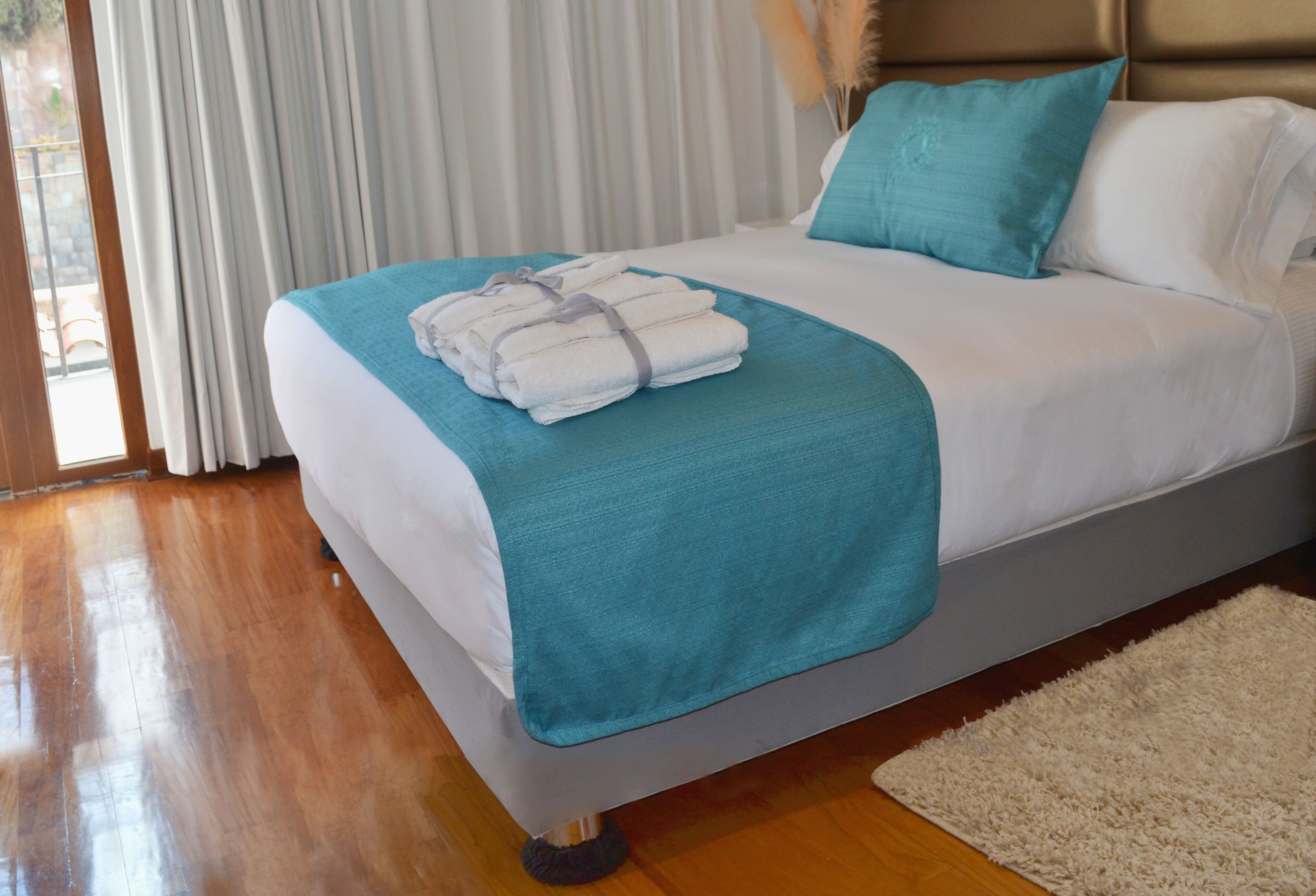 A neatly made bed with a teal throw blanket, white pillows, and a folded white towel on the side.