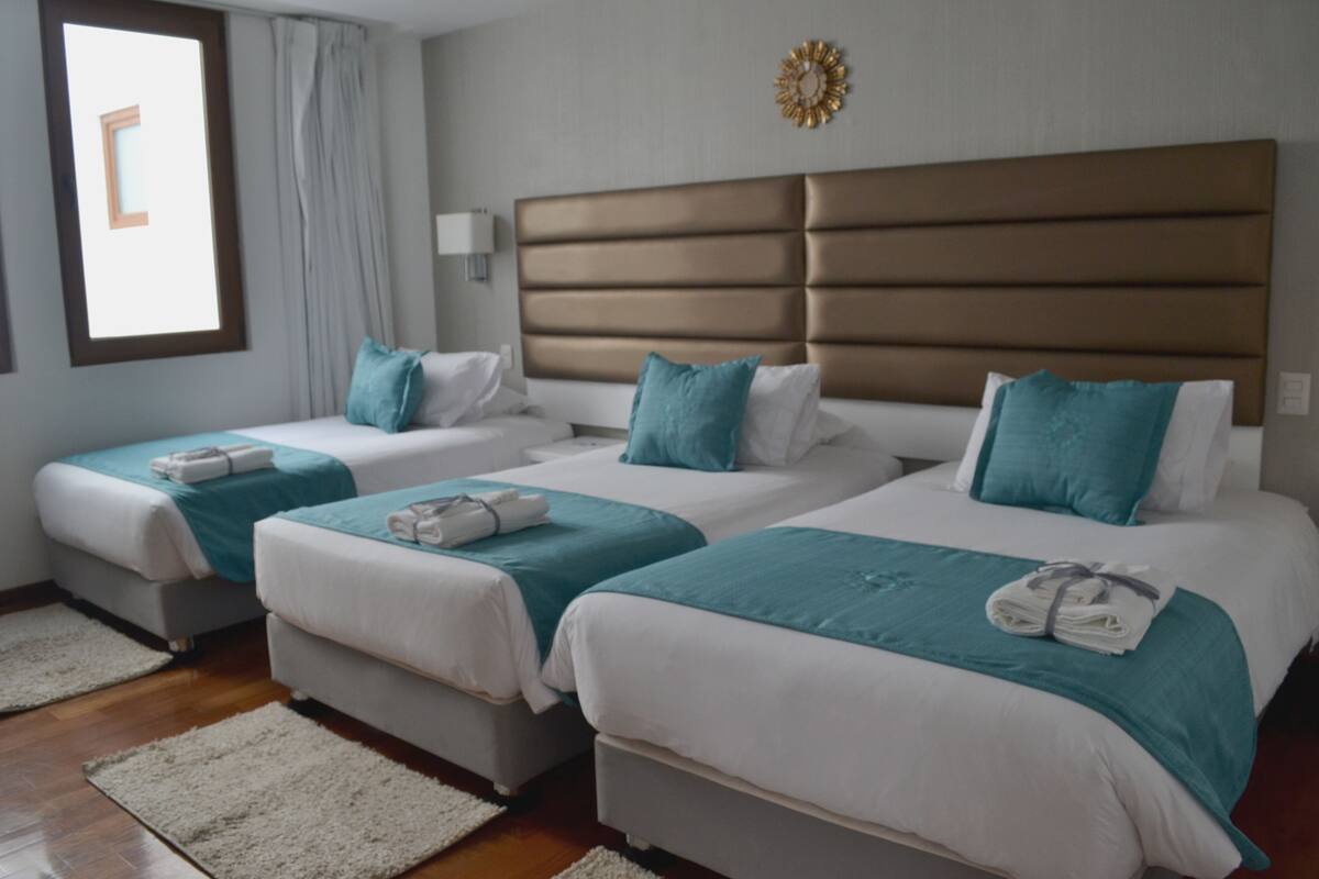 A hotel room with three beds, each with white bedding and teal accents. There is a large window with curtains, a wall-mounted light, and a decorative wall piece.