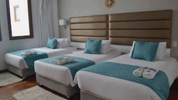 A hotel room with three beds, each with white bedding and teal accents. There is a large window with curtains, a wall-mounted light, and a decorative wall piece.