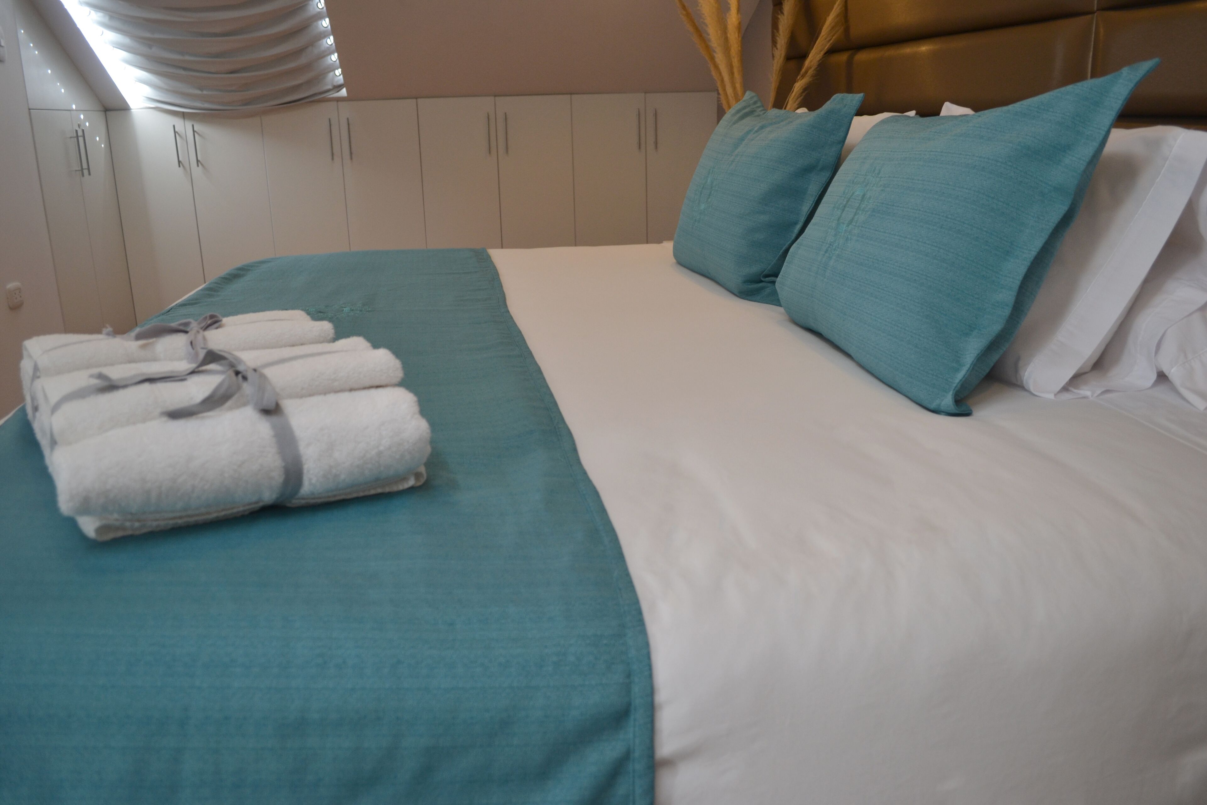 A neatly made bed with teal and white bedding, a stack of white towels, and teal pillows.