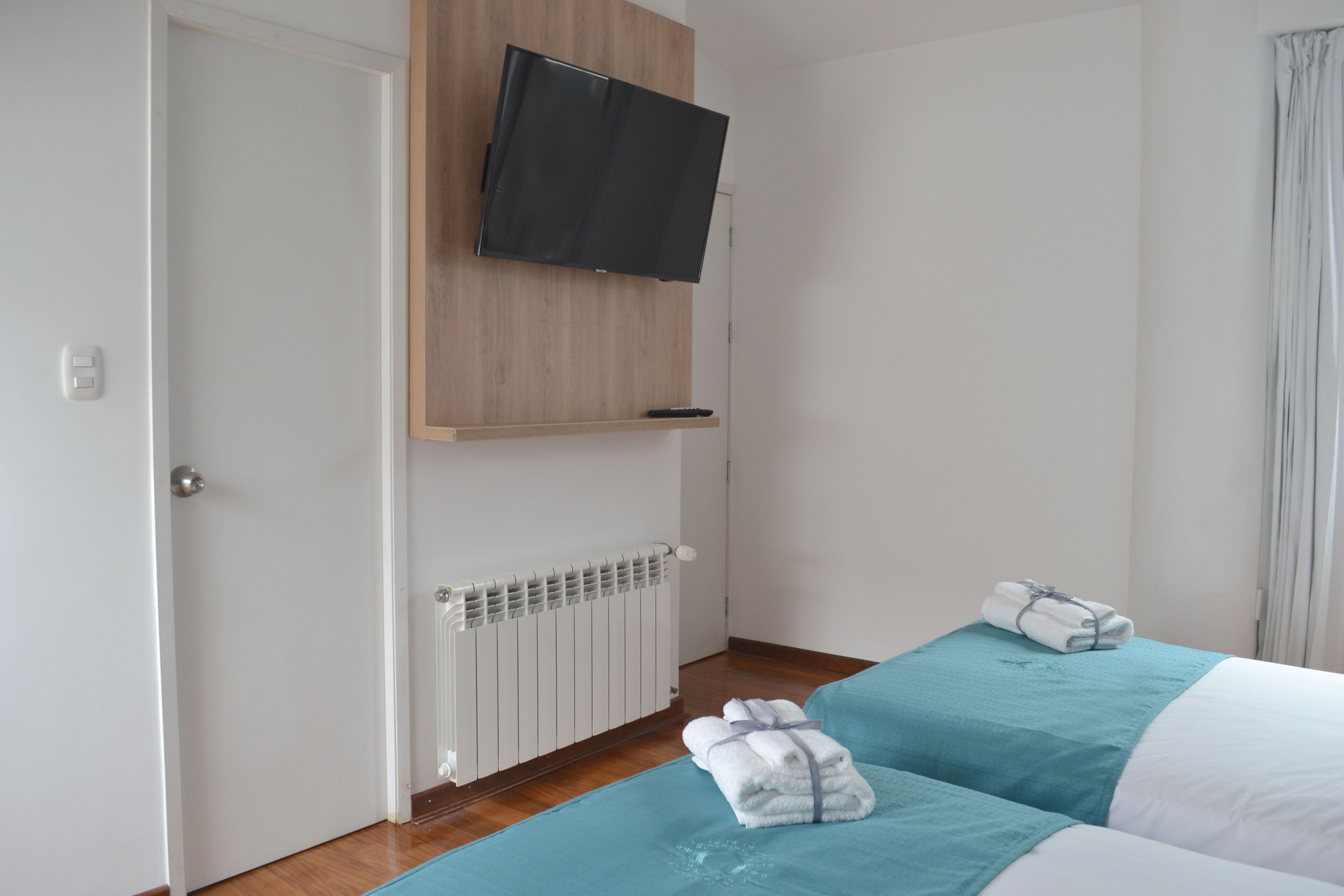 A hotel room with a bed, a TV mounted on a wooden panel, a radiator, and a window with curtains.