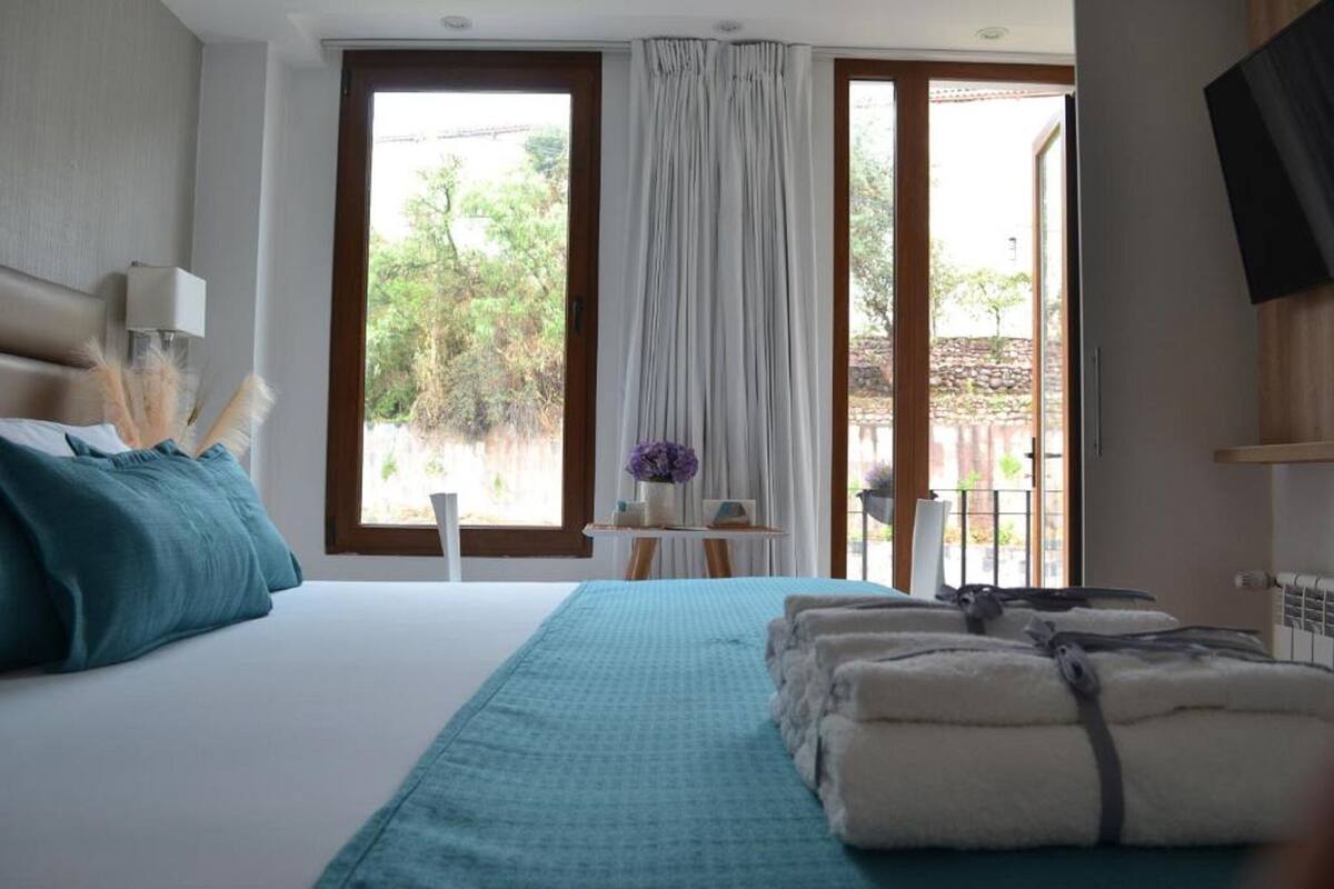 A hotel room with a bed, a small table with a vase of flowers, and a view of the outside.