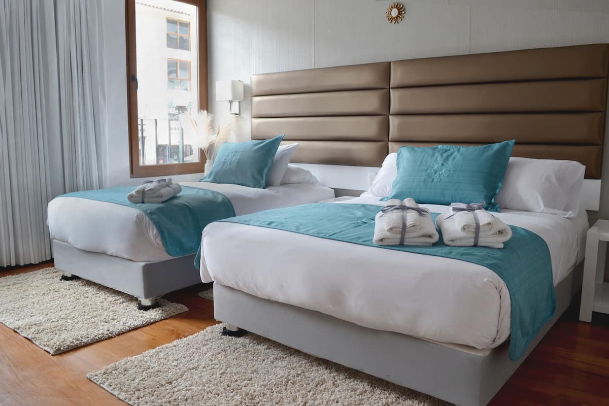 A hotel room with two beds, each with a teal and white bedspread, and a brown headboard. There are white nightstands with lamps, a window with curtains, and a view of buildings outside.