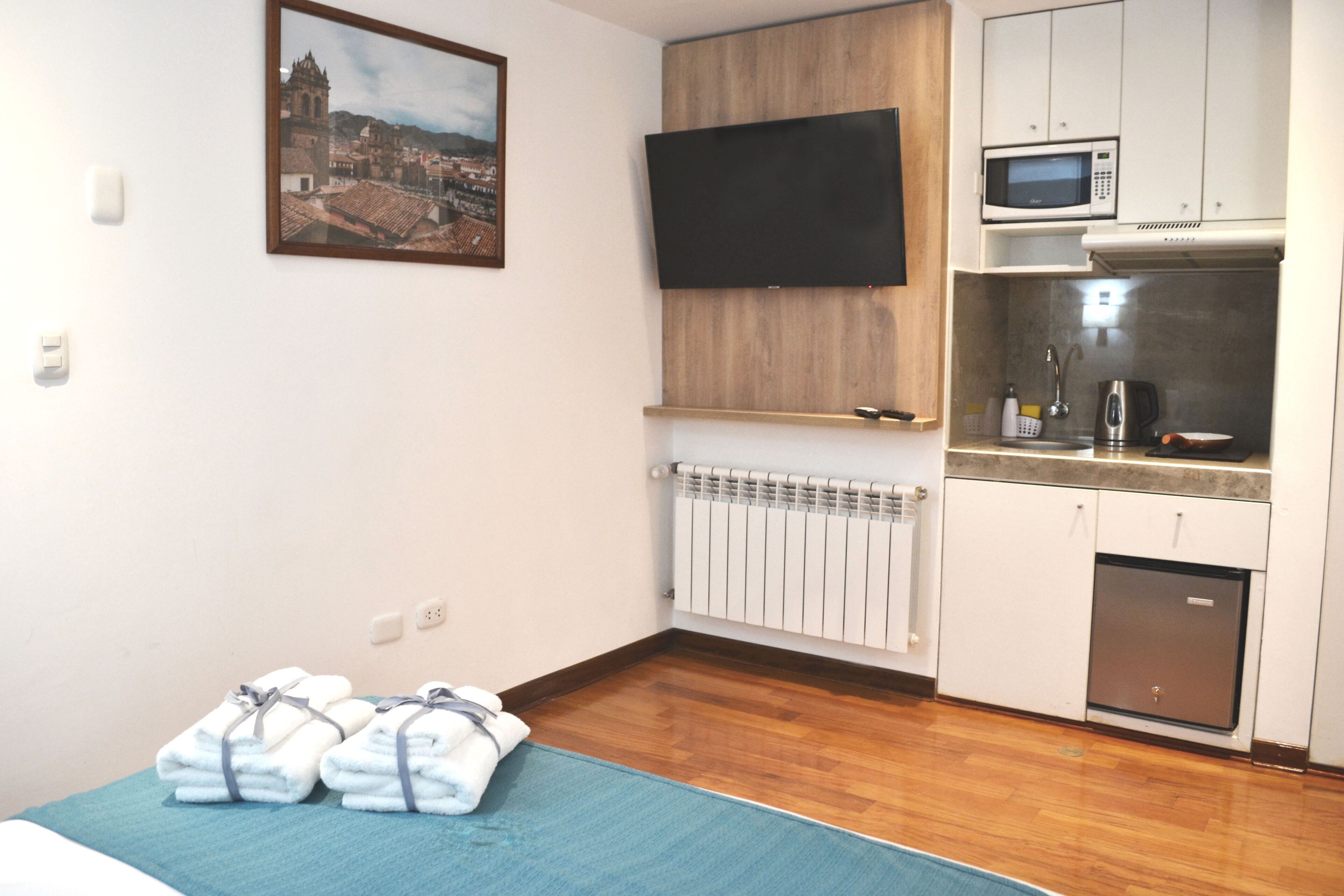 A compact hotel room with a bed, a small kitchenette, a mounted TV, and a cityscape painting on the wall.