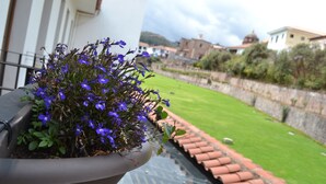 A potted plant with purple flowers on a balcony overlooking a garden with a tiled roof and a stone wall.