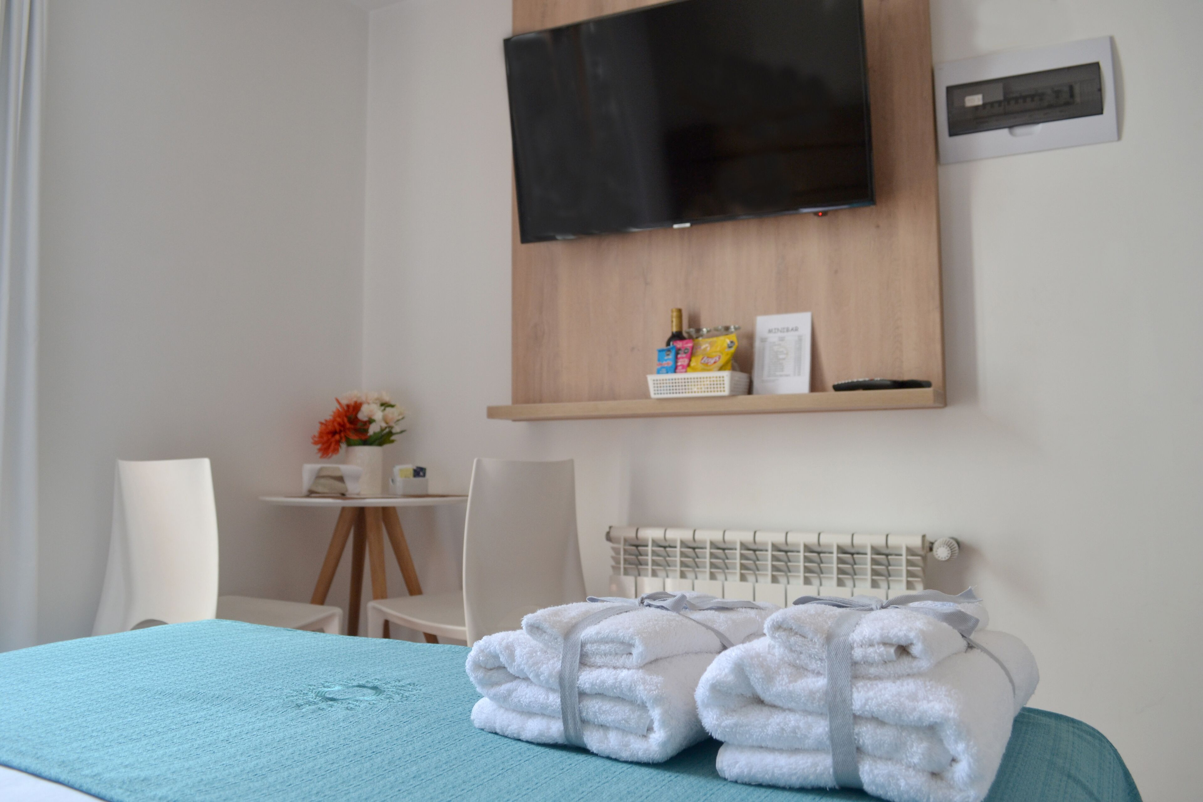A hotel room with a bed, a TV mounted on the wall, a small table with a chair, and a radiator.