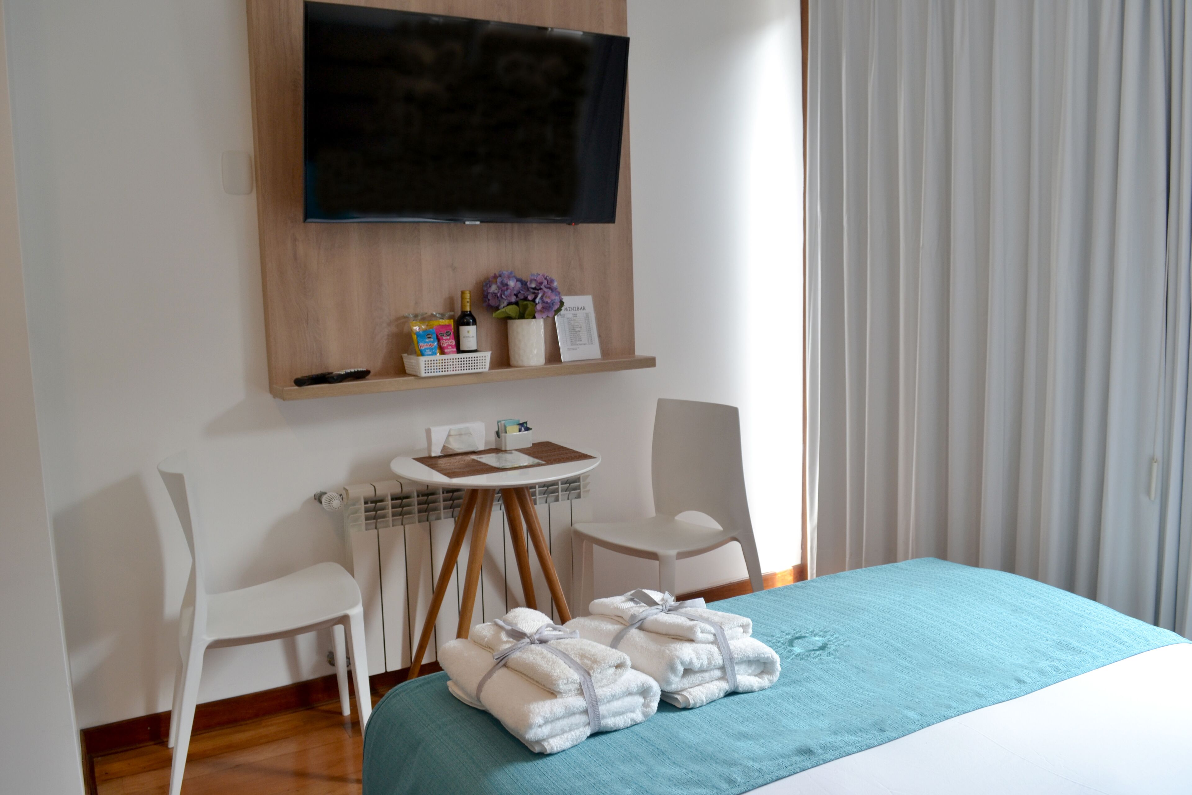 A hotel room with a bed, two chairs, a small table, a TV mounted on the wall, and a window with blinds.