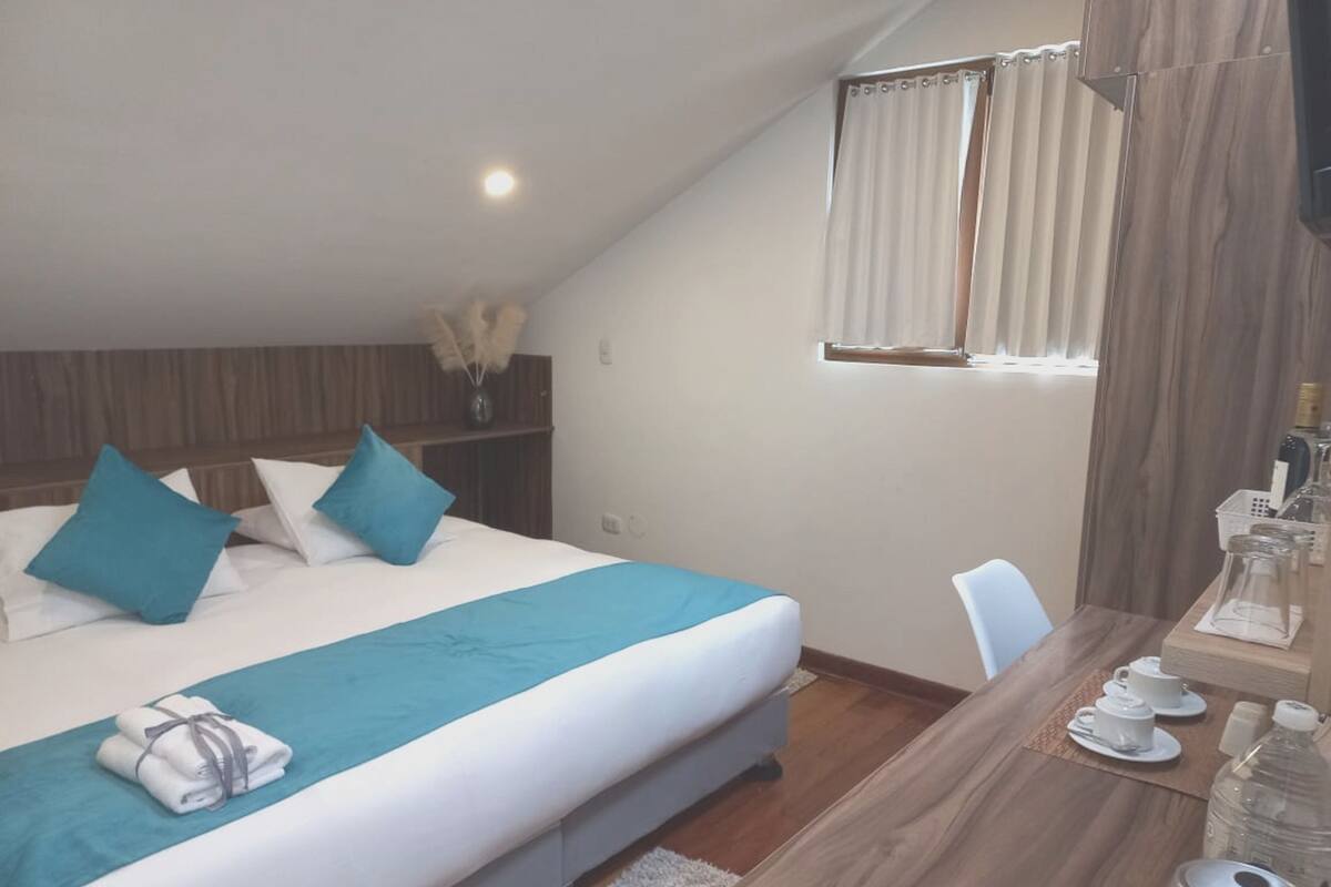 A hotel room with a bed, a desk with a chair, a window with curtains, and a small shelf with decorative items.