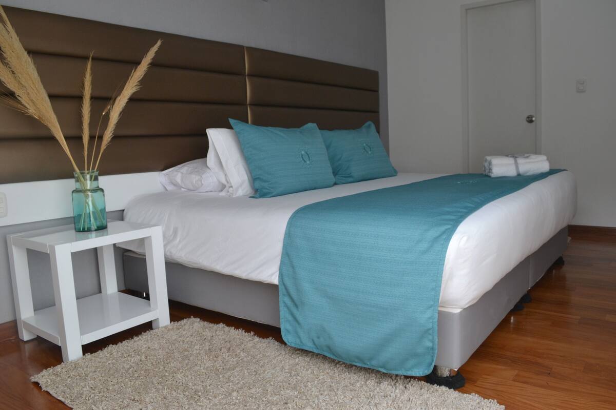 A modern bedroom with a bed, white nightstand, teal bedding, and a decorative vase with pampas grass.