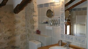 Ein Badezimmer mit Steinwand, Holzbalken, einer weißen Toilette, einem Holzwaschbecken und einem Spiegel.