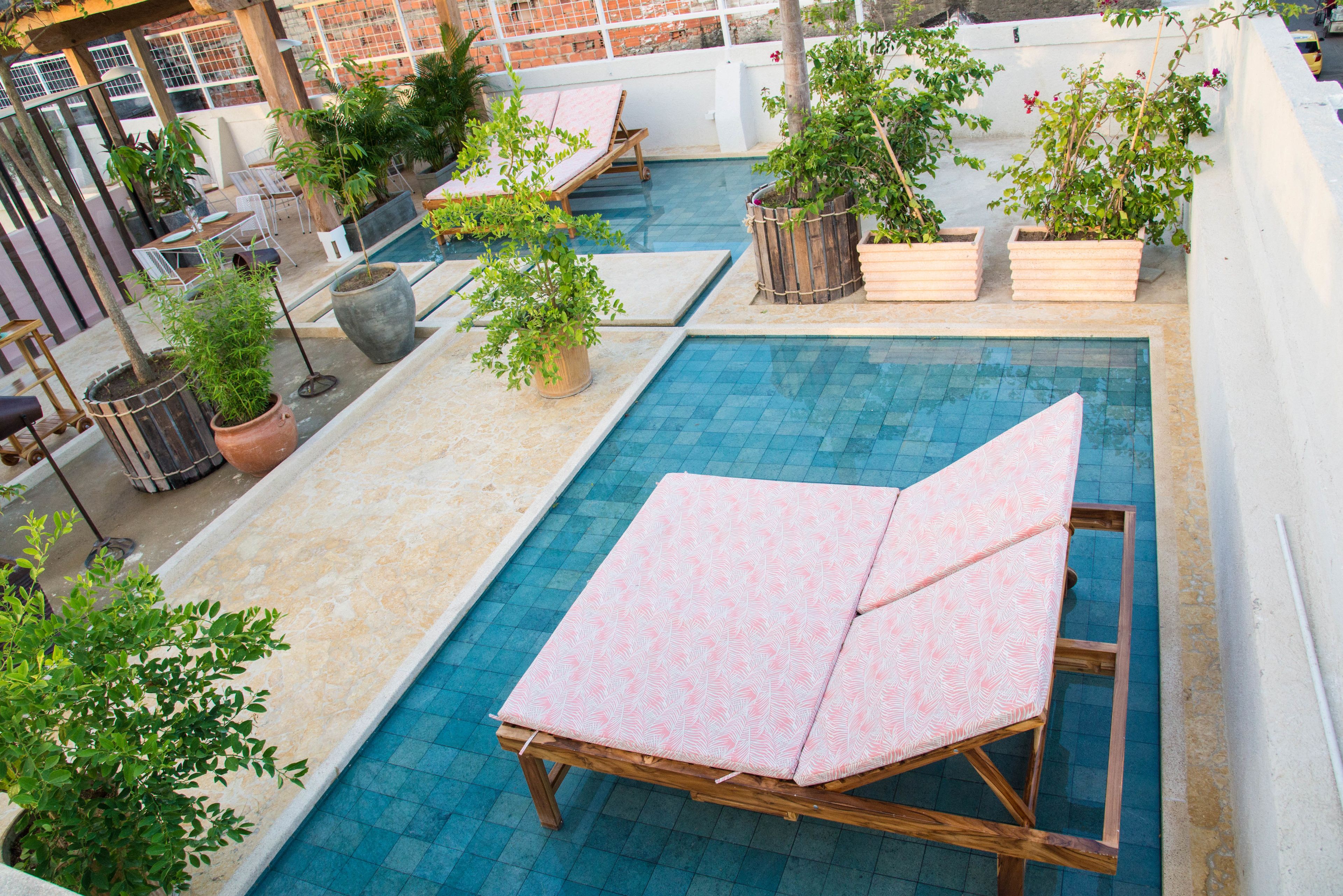 A rooftop pool area with a wooden lounge chair, potted plants, and a tiled pool.