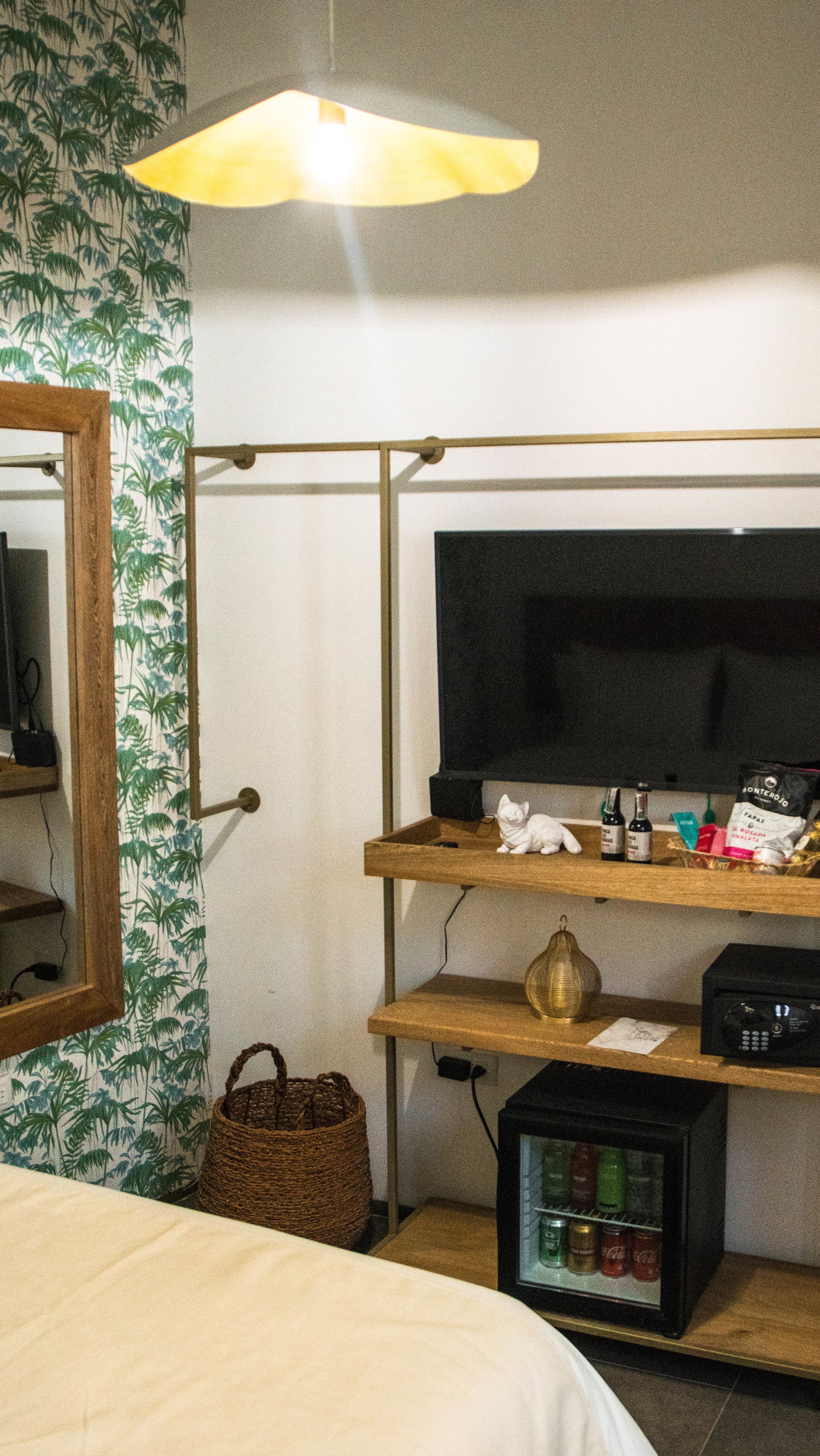 Habitación de hotel con una cama, un televisor sobre un soporte de madera, una mesita con una botella y un bolso, una cesta y un minibar.