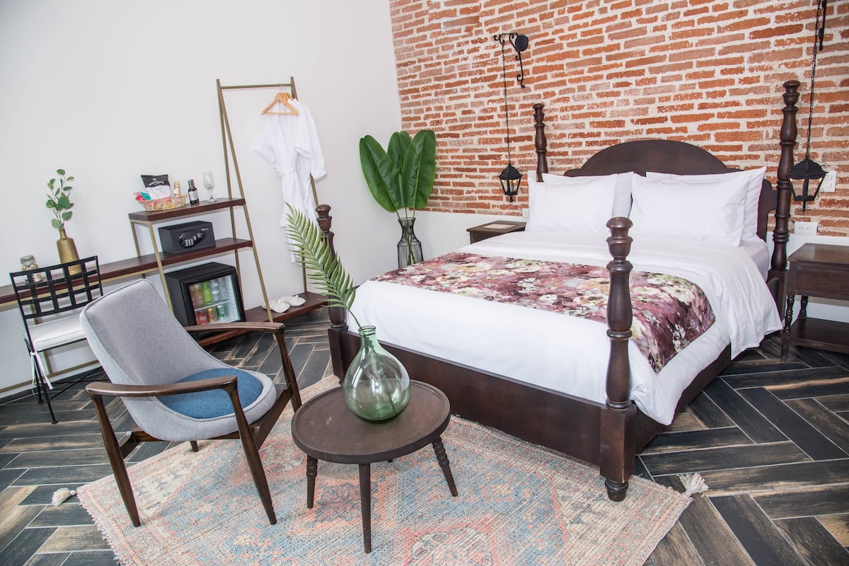 A bedroom with a brick wall, a wooden bed, a chair, a small table, and a shelf with items.