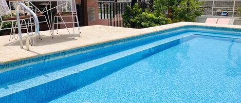 A swimming pool with steps leading into it, surrounded by a wooden deck and white metal chairs.