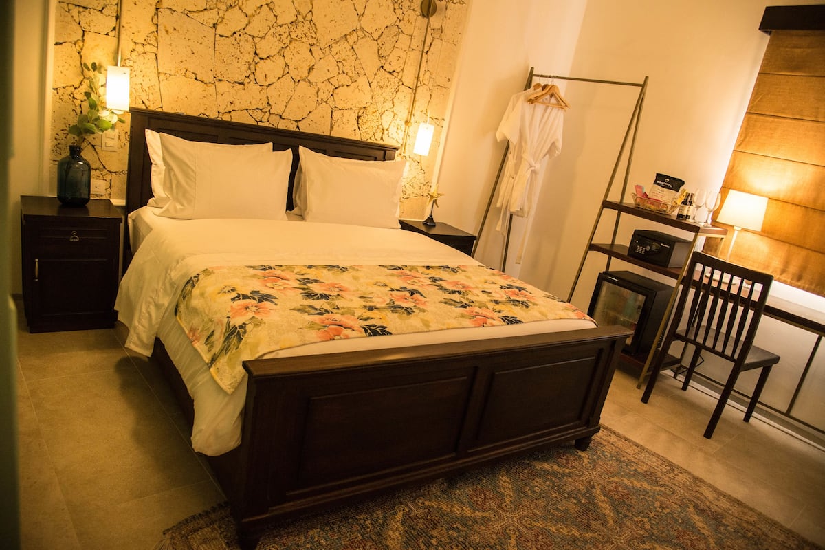 A neatly made bed with a floral bedspread, a bedside table with a lamp, a nightstand, a chair, and a robe hanging on a rack.