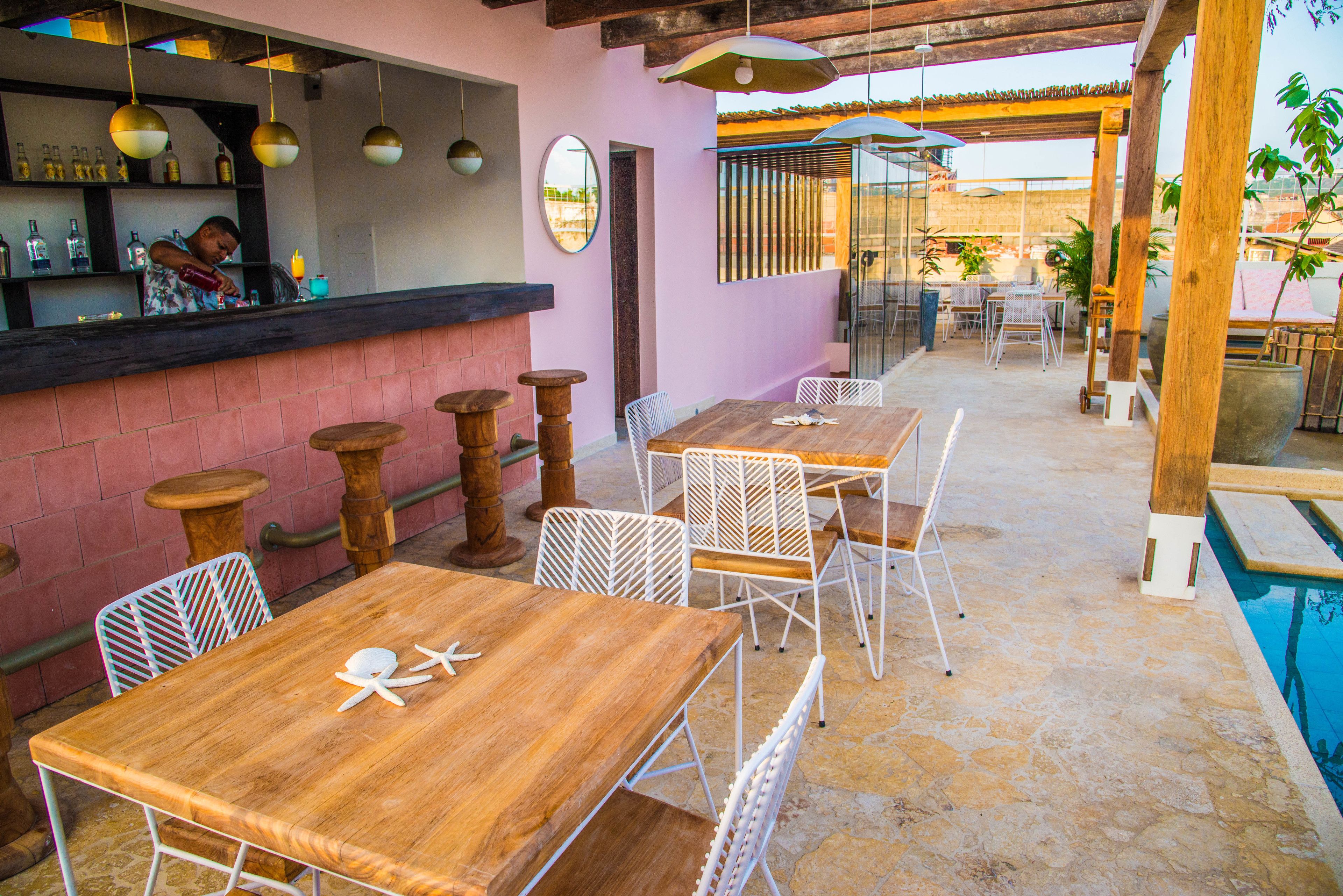 Un'area pranzo all'aperto con tavoli di legno e sedie bianche, un bar con un barista e una piscina sullo sfondo.