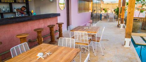 Un'area pranzo all'aperto con tavoli di legno e sedie bianche, un bar con un barista e una piscina sullo sfondo.