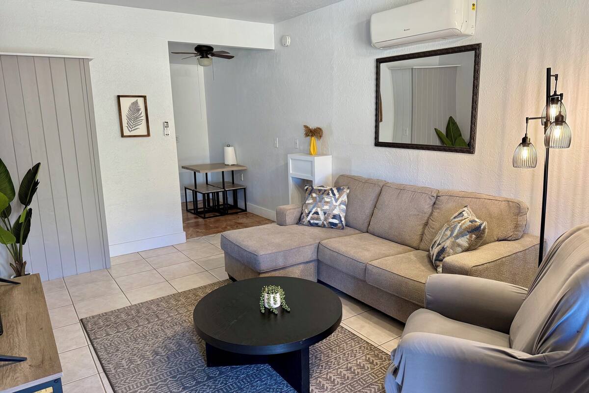 Una sala de estar moderna con un sofá modular, una mesa de centro redonda, un tapete estampado, un comedor con una mesa pequeña y sillas, y un ventilador de techo.