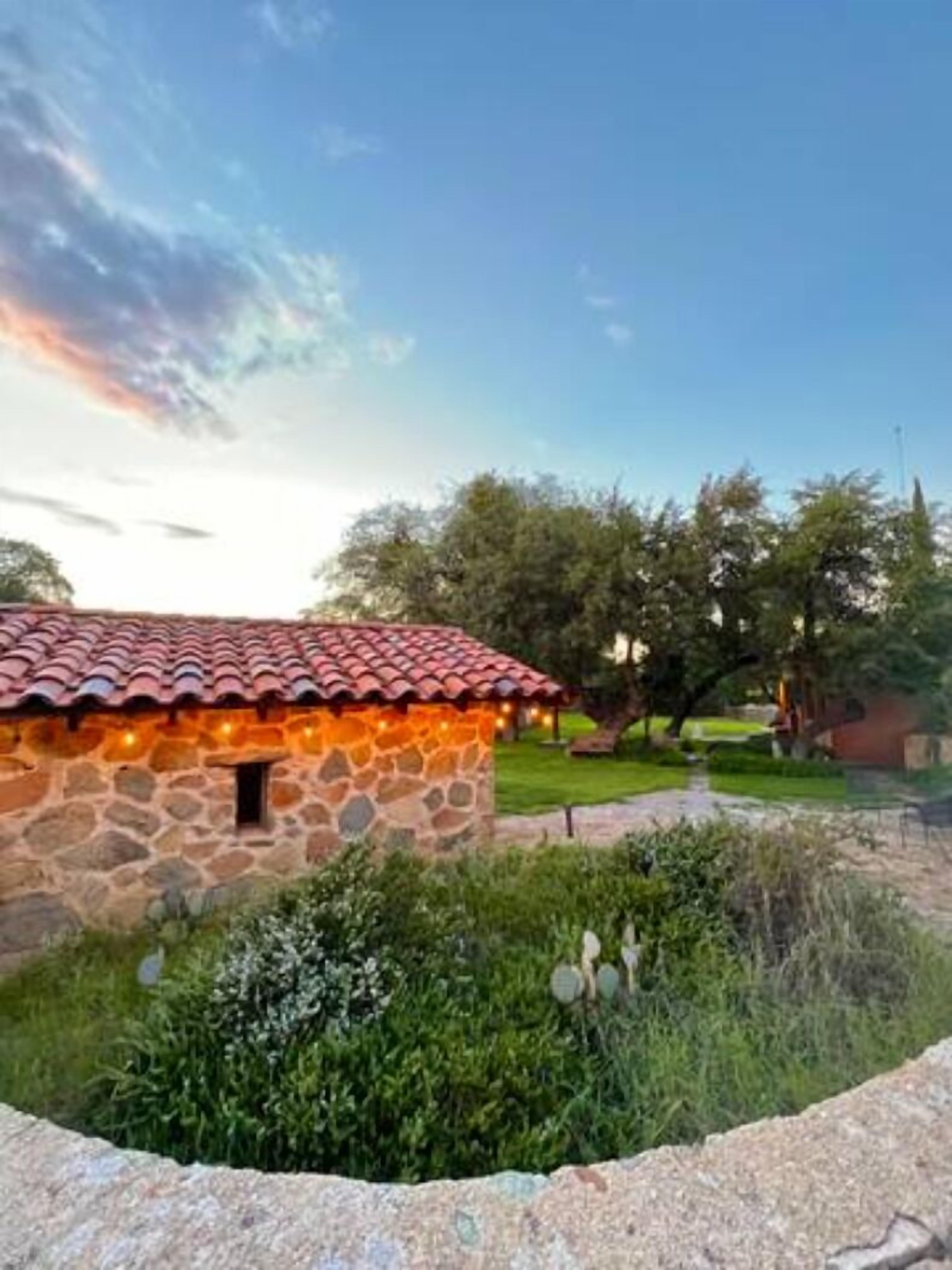 A stone building with a red-tiled roof, surrounded by greenery and trees.