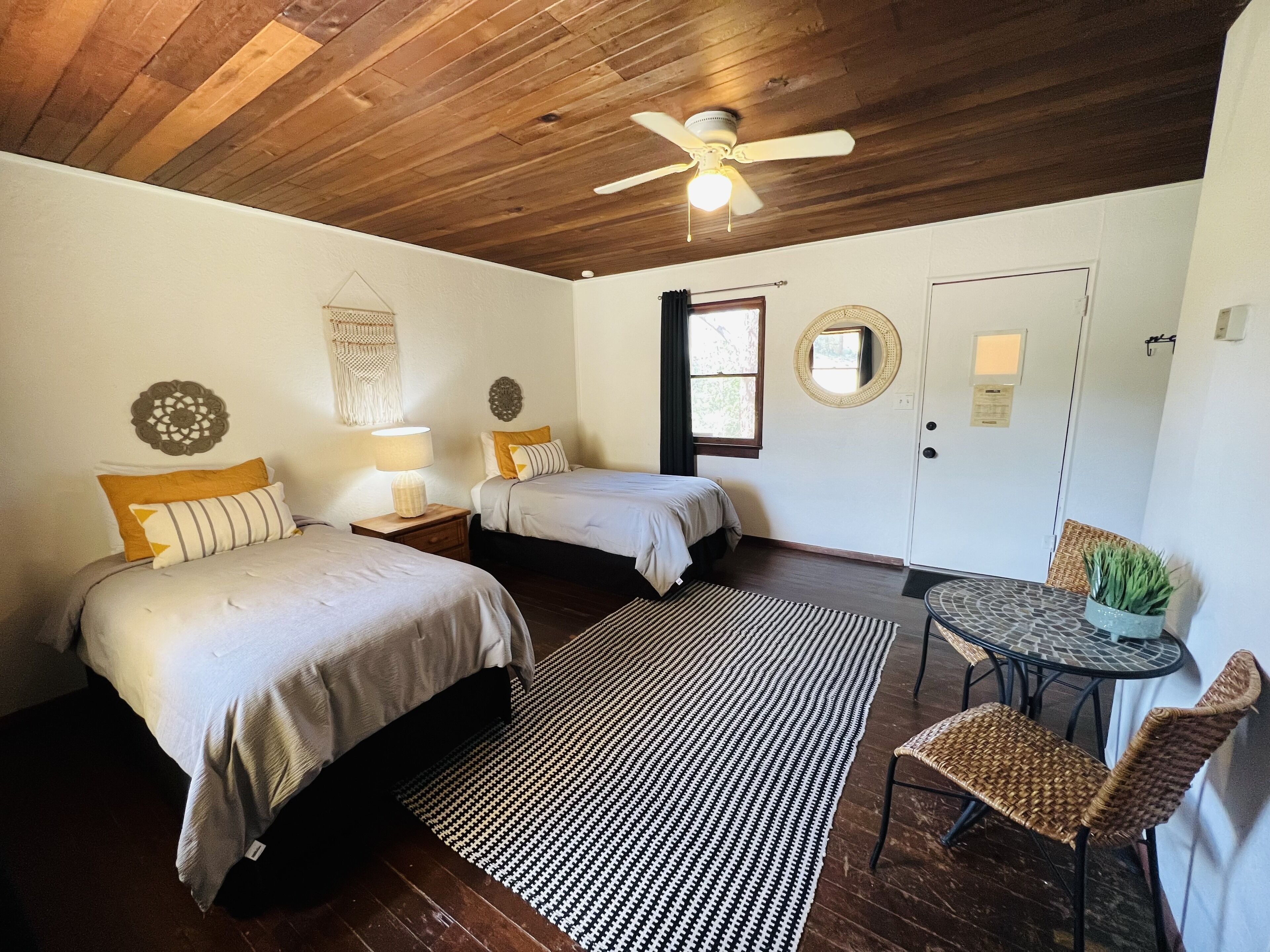 A bedroom with two beds, a ceiling fan, a small table with chairs, and a window with curtains.