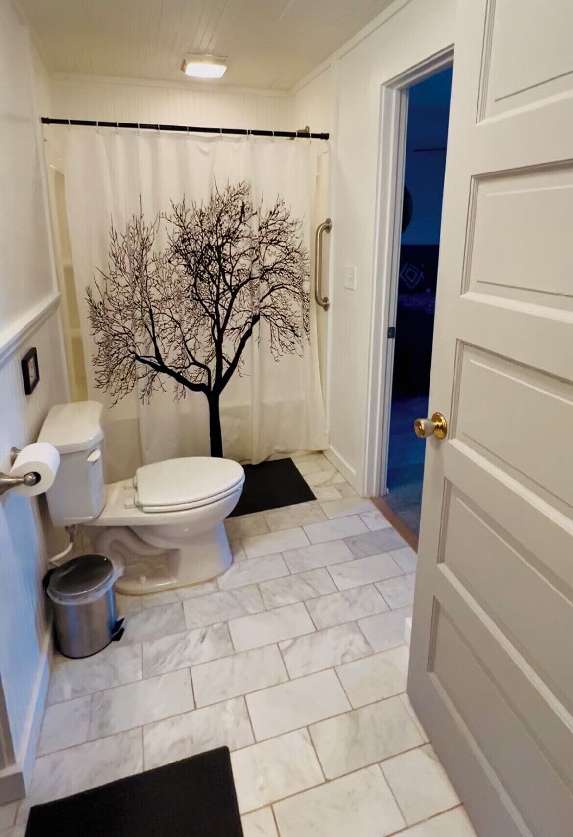 A bathroom with a toilet, a trash bin, a shower curtain with a tree design, and a door leading to another room.