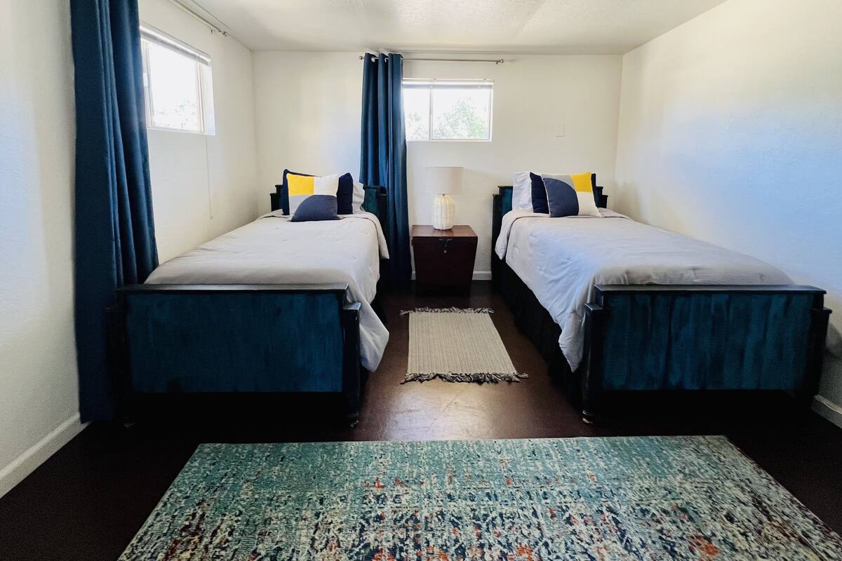 Una habitación pequeña y bien iluminada con dos camas individuales, una mesita de noche con lámpara y una ventana con cortinas.